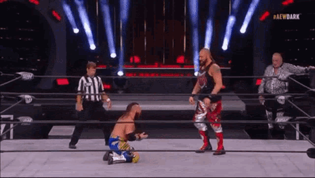 a wrestler is kneeling down in a wrestling ring while another wrestler is standing in the ring .
