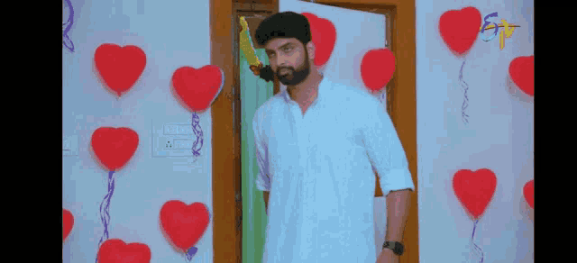 a man with a beard is standing in front of a wall decorated with red hearts and balloons .