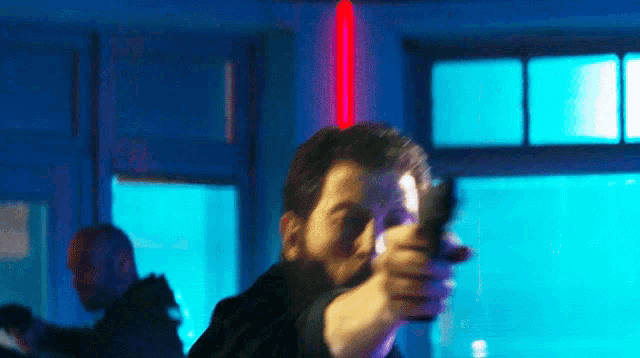 a man with a beard is pointing a gun in a dark room with a red light behind him .