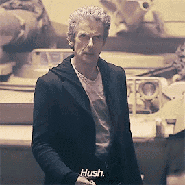 a man in a suit and white shirt is standing in front of a tank and says hush .