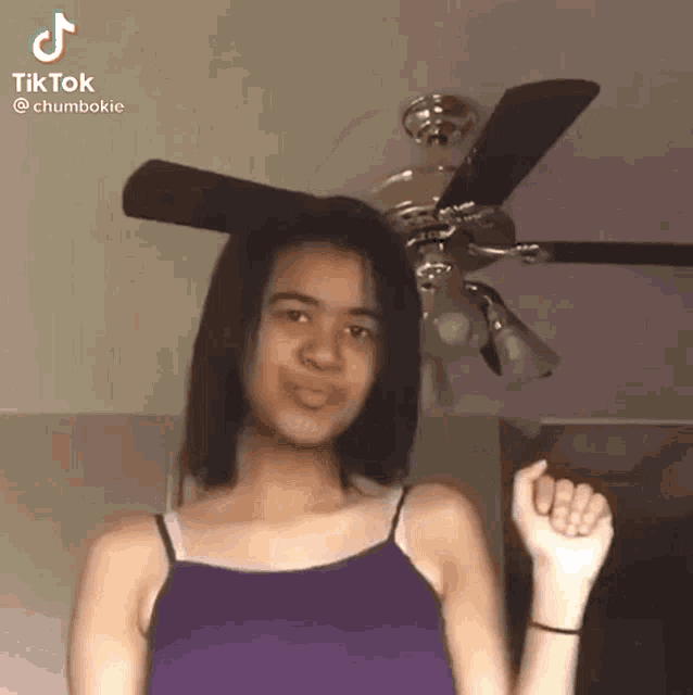 a woman in a purple tank top is standing in front of a ceiling fan and making a funny face .