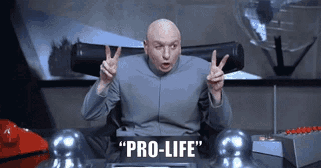 a bald man is sitting at a desk giving a peace sign and says `` pro-life '' .