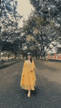 a woman in a yellow dress is walking down a road with trees in the background .