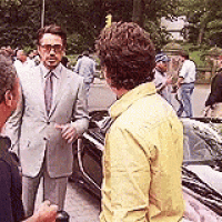 a man in a suit and tie is talking to another man in a yellow jacket while standing next to a car .