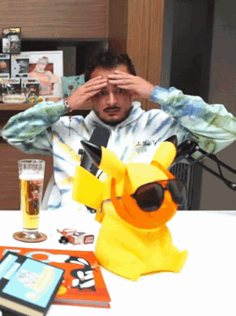 a man wearing a tie dye sweatshirt is sitting at a table with a yellow pikachu wearing sunglasses