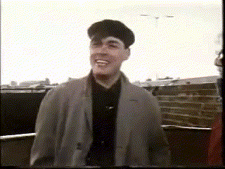 a man in a trench coat and hat is smiling while standing on a balcony .