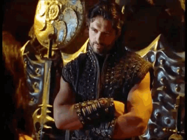 a man with a beard is standing with his arms crossed in front of a throne .