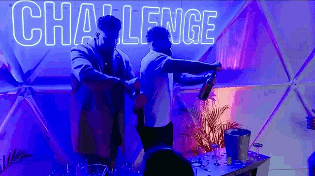 a man is pouring a drink into another man 's hand in front of a neon sign that says challenge .