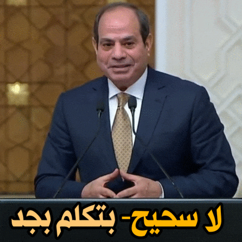 a man in a suit and tie stands at a podium with arabic writing above him