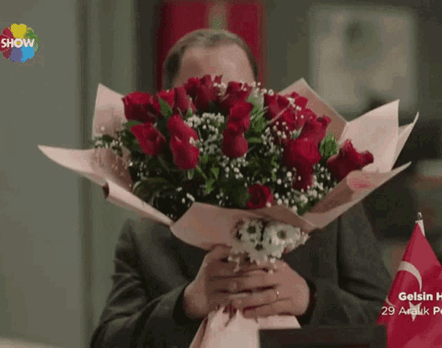 a man is holding a bouquet of red roses in front of his face with a show logo behind him