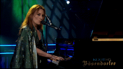 a woman singing into a microphone while playing a piano in front of a sign that says bosendorfer