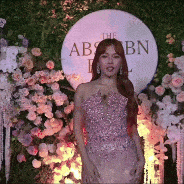 a woman in a pink dress stands in front of flowers and a sign that says the abs cbn