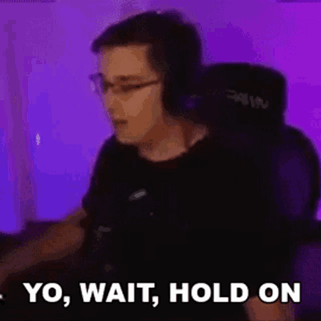 a man wearing headphones and glasses is sitting in a chair with the words `` yo , wait , hold on '' .
