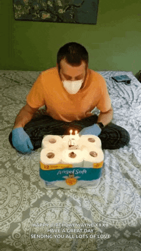 a man wearing a mask and gloves is sitting on a bed with a birthday cake made out of toilet paper rolls