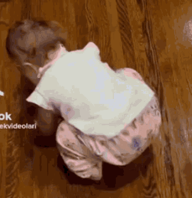 a baby is crawling on a wooden floor in a white shirt and pink pants .