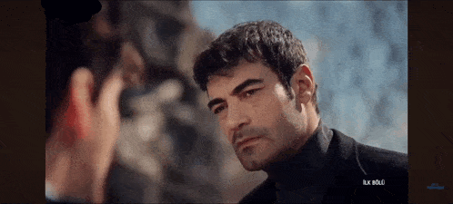 a man in a black turtleneck looks at another man in a black suit