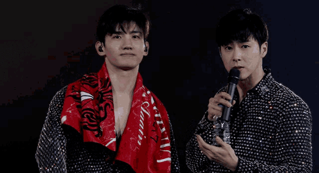 a man holding a microphone stands next to another man with a red scarf around his neck