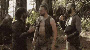 a man without a shirt is talking to two other men in a forest .