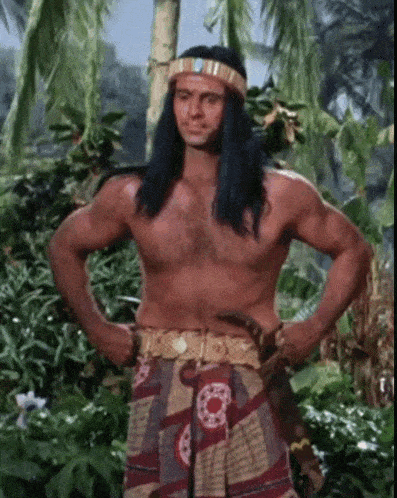 a shirtless man with long hair and a headband is standing in the jungle with his hands on his hips