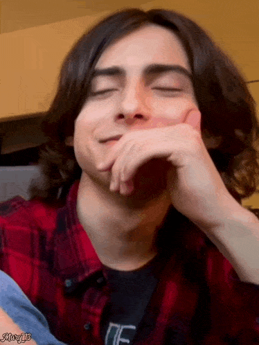 a young man with long hair is wearing a plaid shirt and smiling with his eyes closed .
