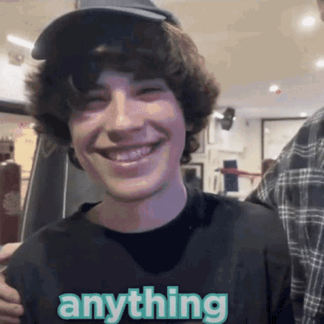 a young man wearing a hat and a black shirt with the word anything on it is smiling .