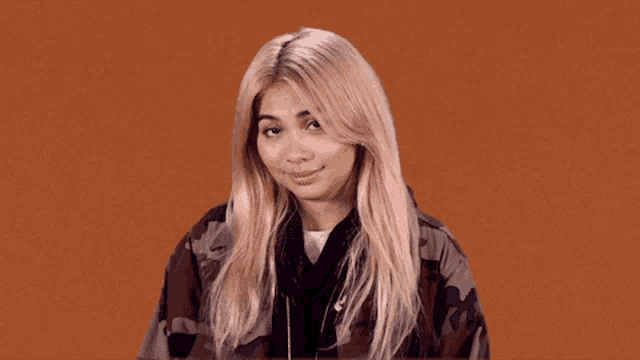 a blonde woman wearing a camouflage jacket looks at the camera