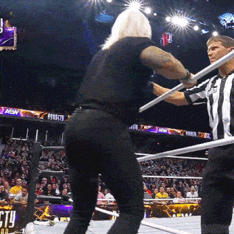 a man in a black shirt is standing in a wrestling ring holding a referee 's baton