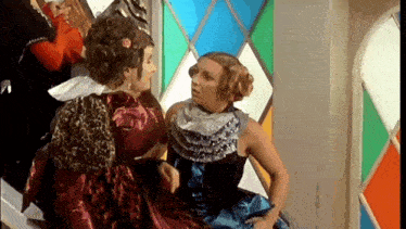 two women in costume are sitting next to each other and talking in front of a stained glass window .