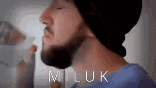 a man with a beard wearing a hat and a blue shirt is drinking from a glass .