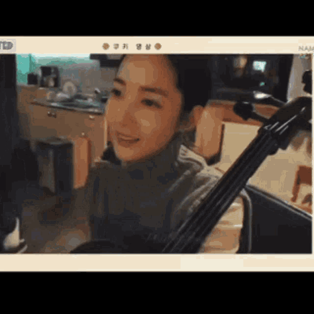 a woman in a grey sweater is holding a cello in front of a kitchen