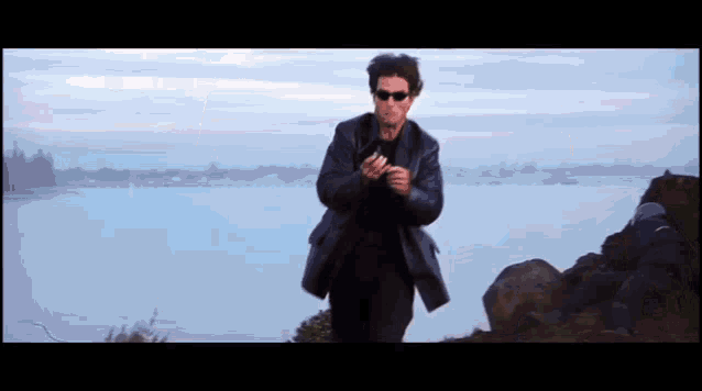 a man in a black jacket and sunglasses is walking on a rocky cliff overlooking a body of water .