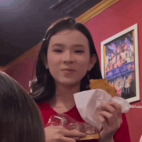 a woman in a red shirt is holding a piece of food in front of a framed picture on the wall .