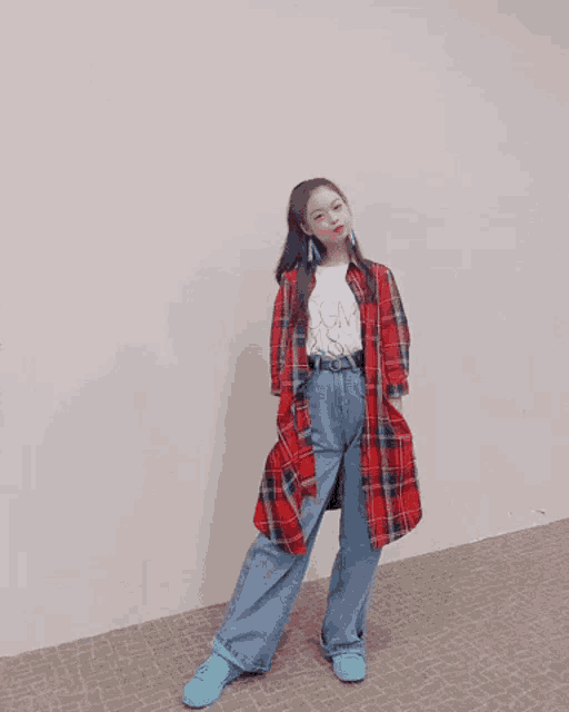 a girl wearing a plaid shirt , wide leg jeans , and blue sneakers is standing in front of a wall .