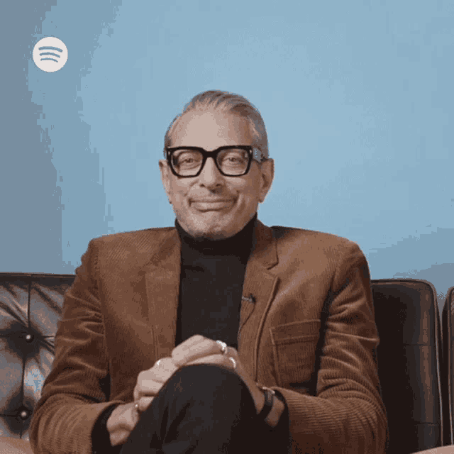 a man wearing glasses and a brown jacket sits on a black couch