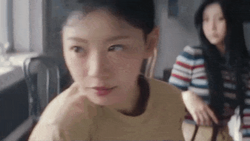 a woman is sitting at a table with her hand on her chin and another woman is sitting behind her .