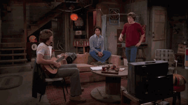 a man in a red t-shirt is standing in a living room with two other men