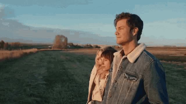 a man and a woman are standing in a field . the man is wearing a denim jacket .