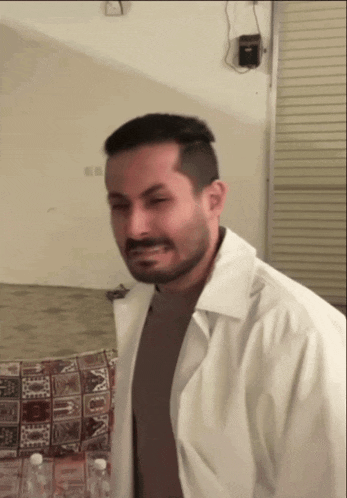 a man in a white coat stands in a room with a bottle of water behind him