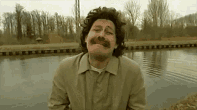 a man with a moustache is standing in front of a body of water .|833x465.959375