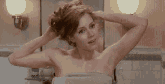 a woman is wrapped in a towel in a bathroom and looking at her hair in the mirror .