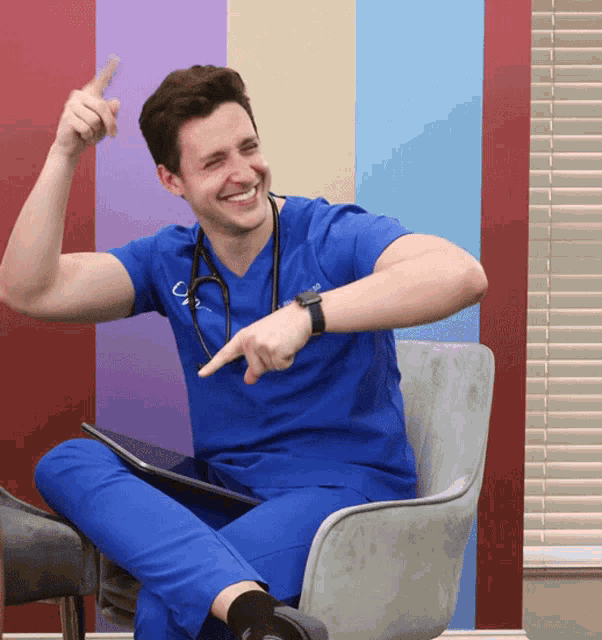 a man in a blue scrub top with a stethoscope around his neck is smiling and pointing