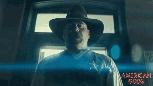 a man in a sheriff 's uniform stands in front of a door with the words american gods above him