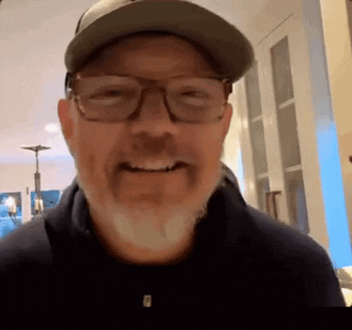 a man with a beard wearing glasses and a baseball cap is smiling .