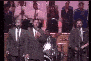 a group of men in suits and ties are singing into microphones on a stage in front of a choir .