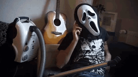 a person wearing a scream mask sits on a couch next to a vacuum cleaner