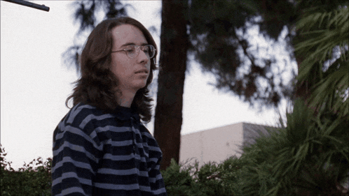 a young man wearing glasses and a striped sweater stands in front of a tree