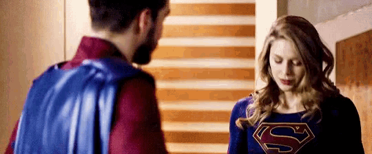 a man and a woman are standing next to each other in a room . the woman is wearing a superman costume .