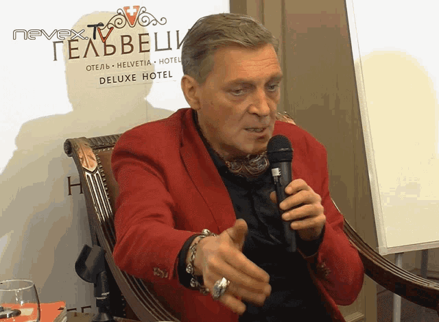 a man in a red jacket holds a microphone in front of a sign that says nevex deluxe hotel