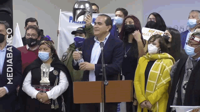 a man stands at a podium surrounded by people wearing masks and holding a sign that says mejor mal