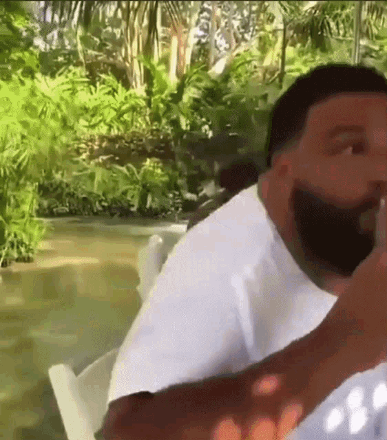 a man with a beard is sitting in a white chair in front of a lush green forest .